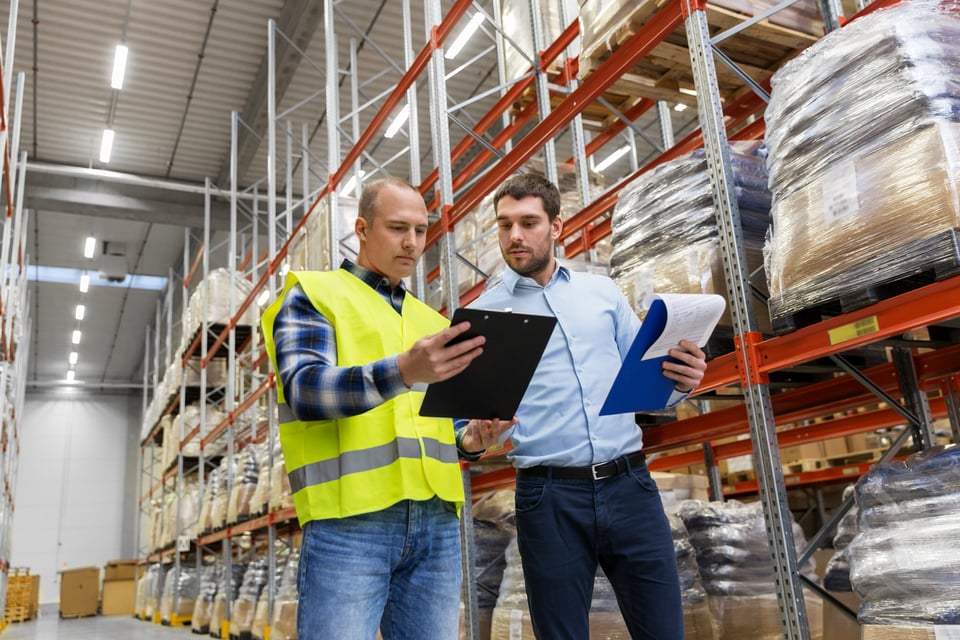 Procurement professional in business attire negotiates product prices with supplier wearing yellow warehouse vest.