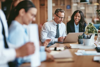 Businessmen and businesswomen sit at conference table, reviewing corporate supplier diversity spend on laptop computer.