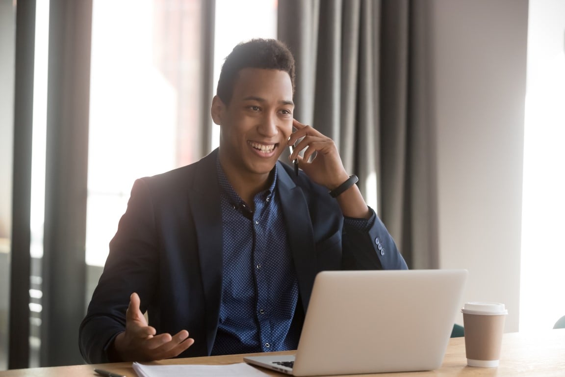 Salesman sits at desk, on which rests his laptop computer, while he speaks with customer on smartphone.  