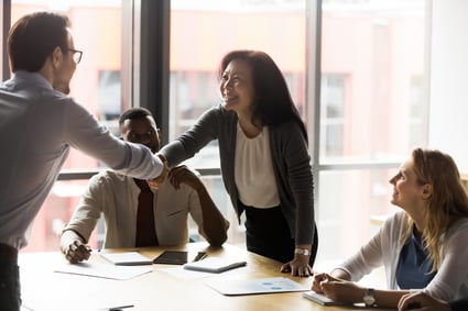 Female Korean business owner stands to shake hands with white man as Black male and white female colleagues watch.  