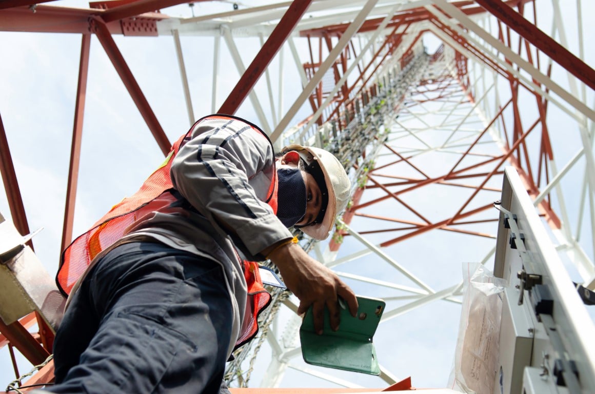 Maintenance worker stands beneath tall cellular phone tower, reviewing data on his smartphone. 