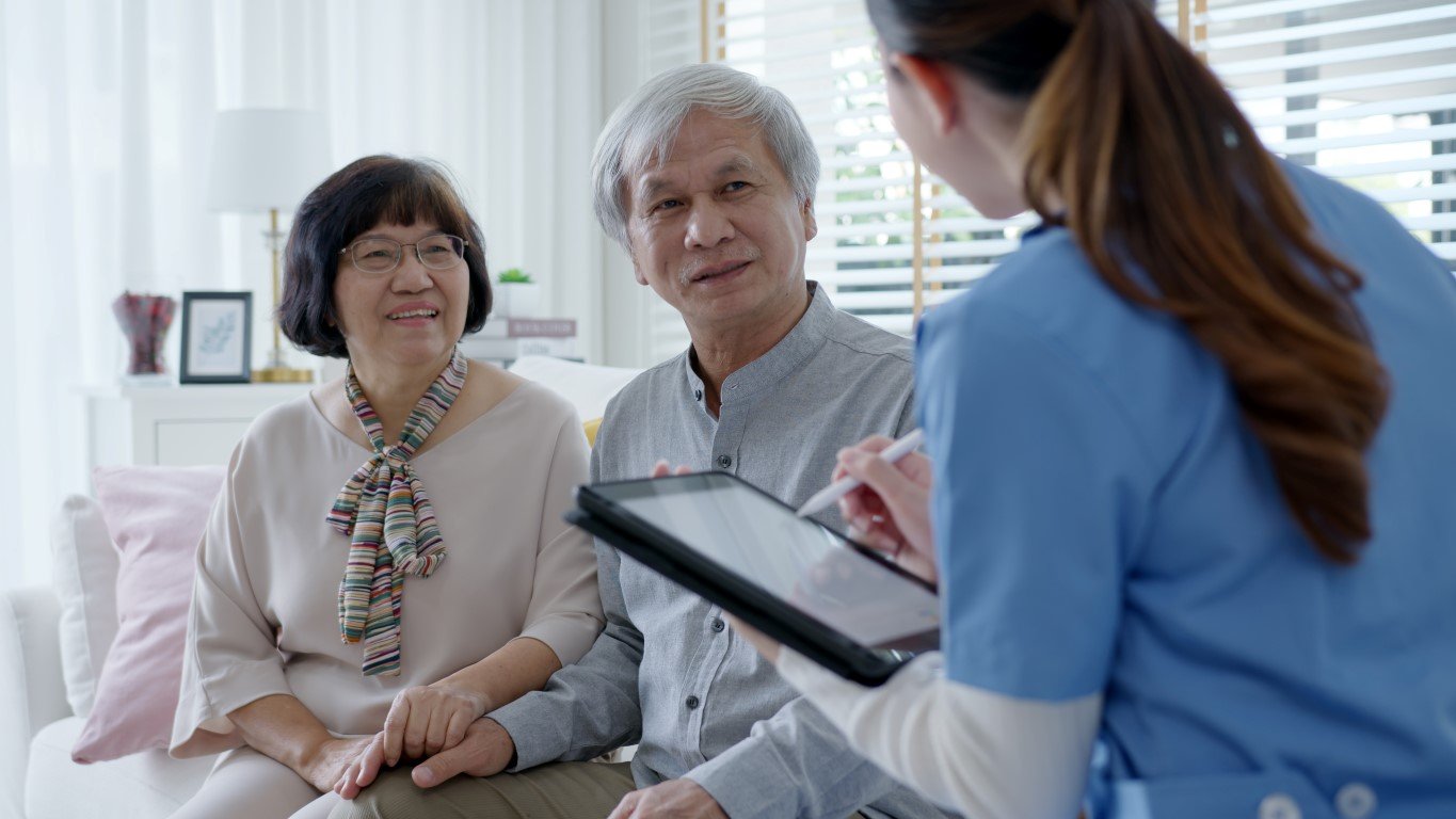 Home health care worker speaks with senior couple in their home, recording their data with a stylus on her tablet computer. 