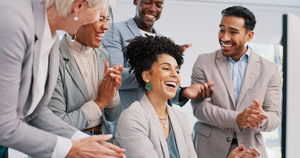 Procurement manager’s colleagues stand around her and applaud as she displays cost savings on her computer screen.