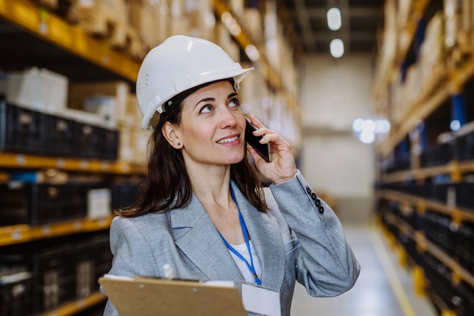 Manager stands in warehouse, wearing hard hat, talking on her cell phone with suppliers about procurement inflation strategies.