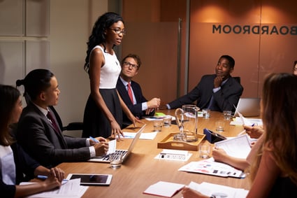 Black, Asian, and white members of a corporate board sit around conference table listening to female business owner speaking. 