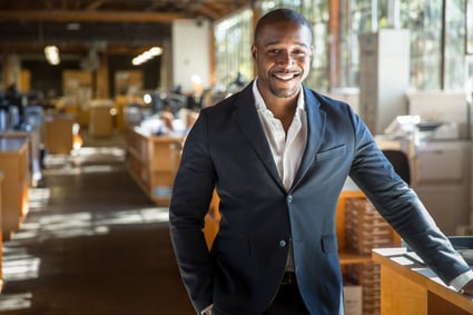 Black business owner stands in office, desks and filing cabinets visible in background.