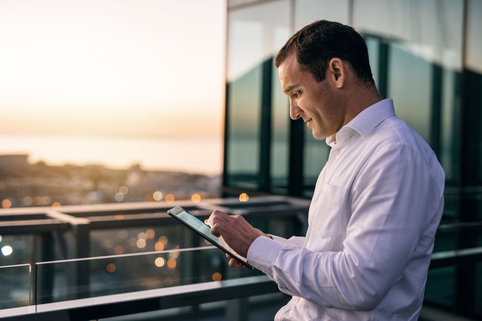 Manager stands on office building balcony, using tablet computer to review success of his procurement inflation strategies. 